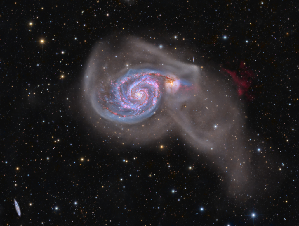 Mitten im Bild leuchtet eine Spirale in vielen Farben, die von einem verschwommenen Nebel umgeben ist. In derl inken unteren Ecke ist eine kleine Galaxie von der Seite zu sehen.