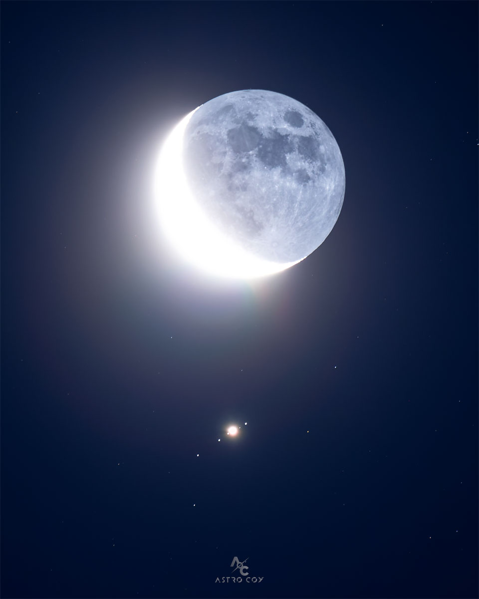 Der Erdmond in der Sichelphase befindet sich über der Bildmitte. Gleich darunter leuchtet ein heller Fleck, der von vier weiteren Lichtpunkten in einer Reihe umgeben ist, die allesamt Jupiters Monde sind.