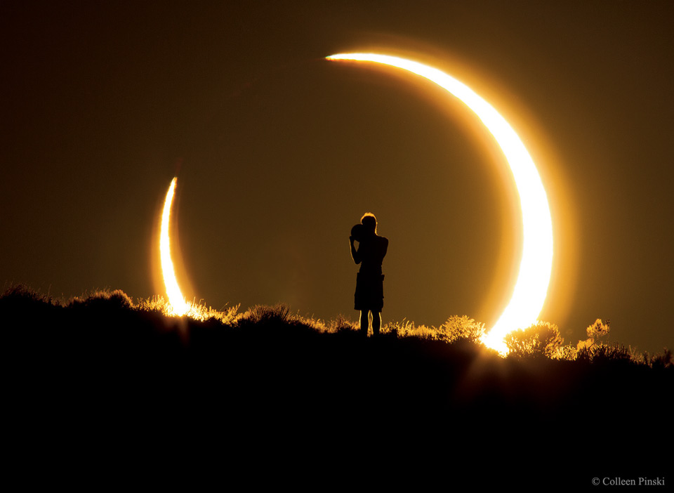 Über dem Horizont ist eine riesige Sonnensichel zu sehen, die entsteht, weil der Mond teilweise die Sonne bedeckt. Vor der Sonnenfinsternis steht eine Person.