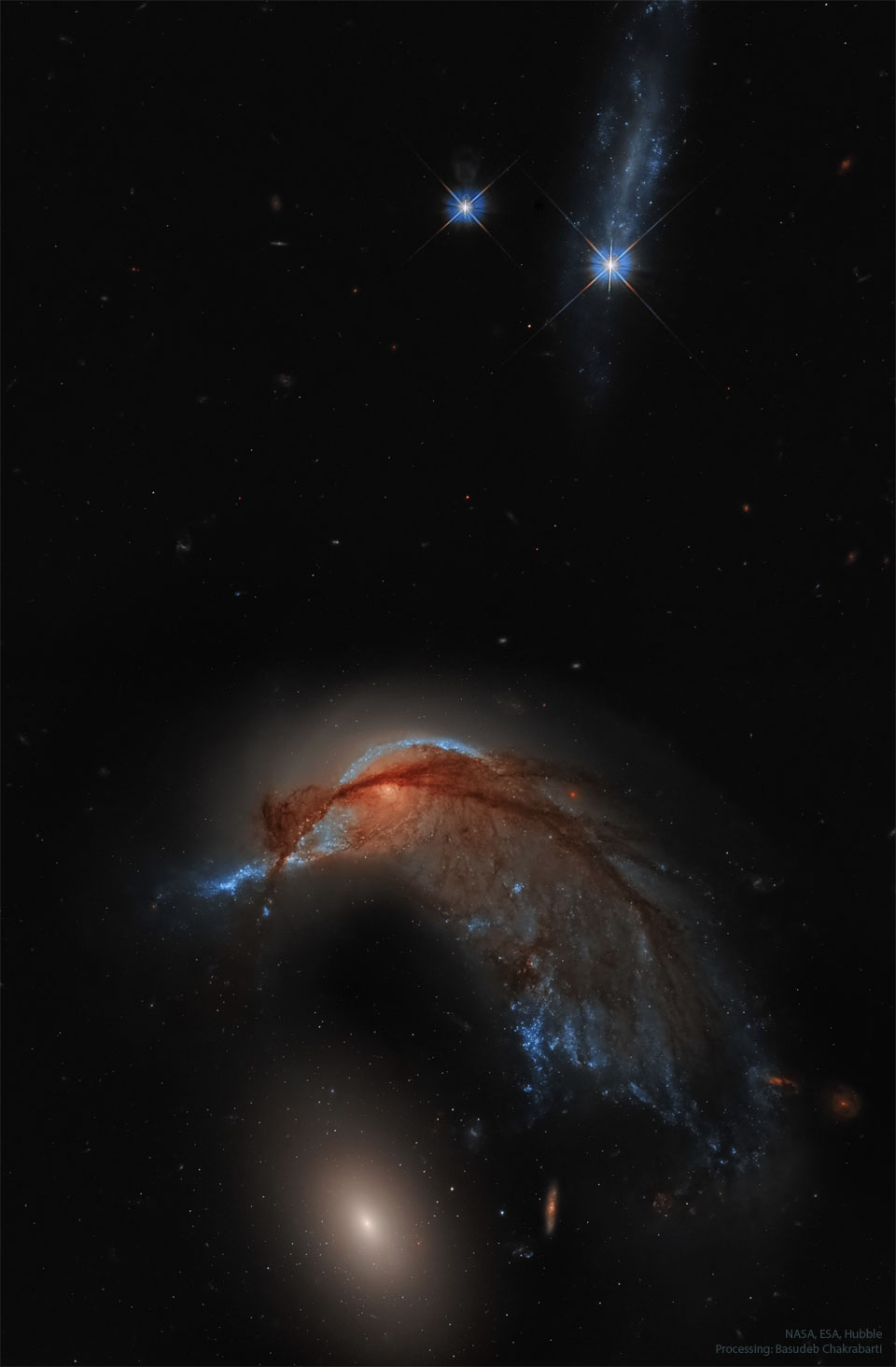 Oben im Bild leuchten zwei gezackte blaue Sterne. Untn ist eine aufgezogene, stark verzerrte Galaxie mit vielen dunklen Fasern und Staubbahnen, darunter befindet sich eine strukturlos wirkende elliptische Galaxie. Der Hintergrund des Bildes ist dunkel und wirkt auf den ersten Blick sternenlos.