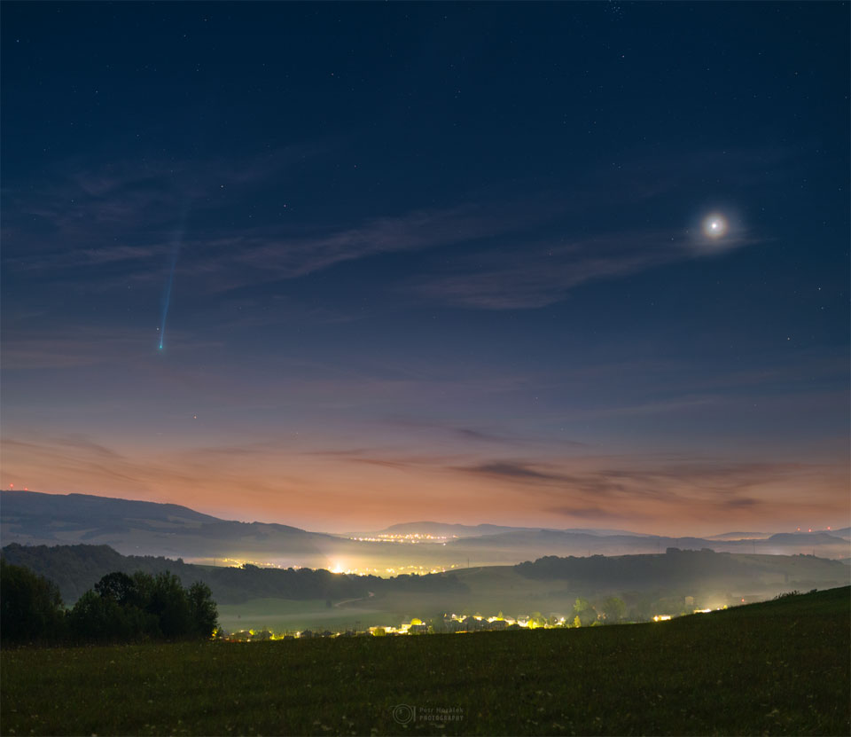 Malerische, hügelige Landschaft kurz vor Sonnenaufgang. Links steht der Komet Nishimura in der Nähe des Horizonts mit einem langen Schweif, der zum oberen Bildrand hin abnimmt. Der helle Fleck rechts ist die Venus. Der Himmel bei Sonnenaufgang ist oben dunkelblau und geht am Horizont in hellbraun über, die Hügel im Vordergrund sind grün.