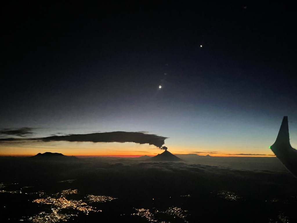 Über einer dunklen Landschaft mit den Lichtern einer Stadt zeichnet sich vor der Dämmerung am Horizont die Silhouette eines Vulkans mit Rauchfahne ab. Darüber leuchten am dunklen Himmel der Mond und die Venus.