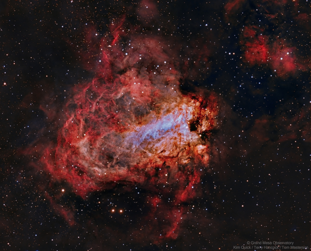 Mitten im Bild leuchtet ein roter, verwirbelter Nebel mit dunklen Strukturen, mit einem hellblauen Bereich in der Mitte.