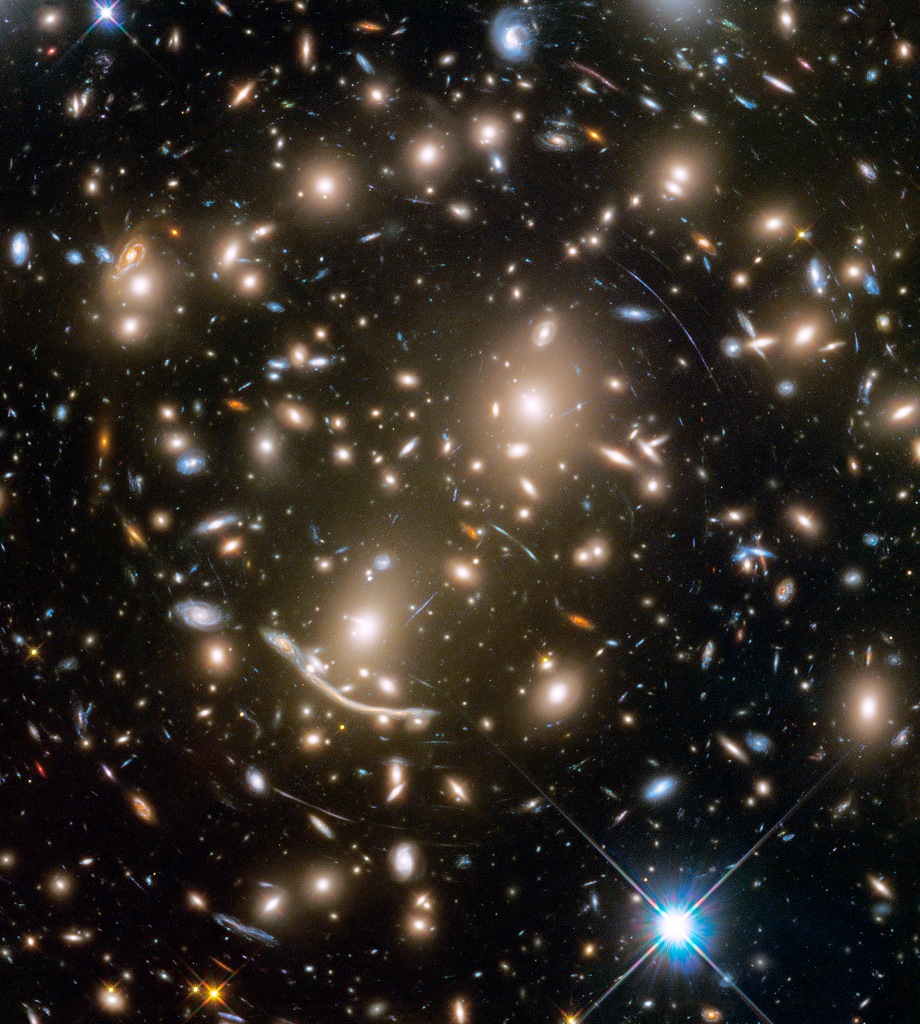 Das Bild ist voller Galaxien. Im Vordergrund befinden sich unverzerrte Galaxien, dazwischen sind schmale Bögen von weit dahinter liegenden, stark verzerrten Galaxien verteilt.