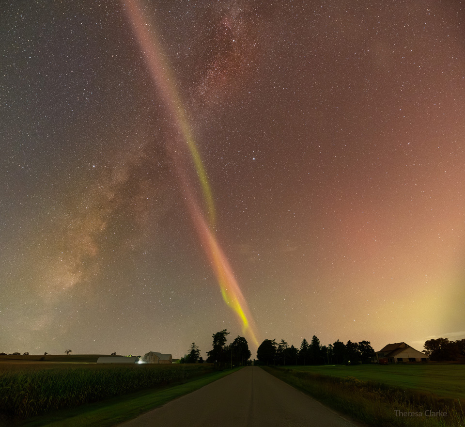 Über einem Horizont mit Bäumen steigt diagonal nach rechts die Milchstraße auf und diagonal nach links ein Lichtband, ein sogenannter STEVE. Beide kreuzen sich und bilden ein X.