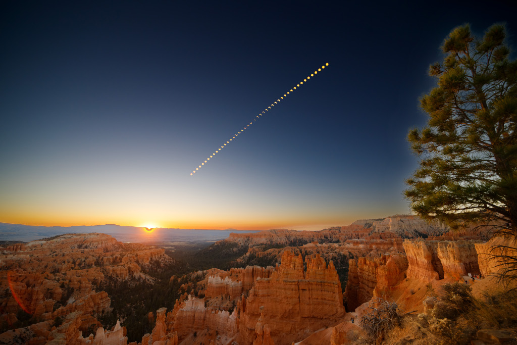 Im Bryce Canyon geht am Horizont links neben der Mitte die Sonne auf. Am dunkelblauen Himmel verläuft diagonal die Sonne, die zunehmend vom Mond verdeckt wird und dann wieder hervorkommt.
