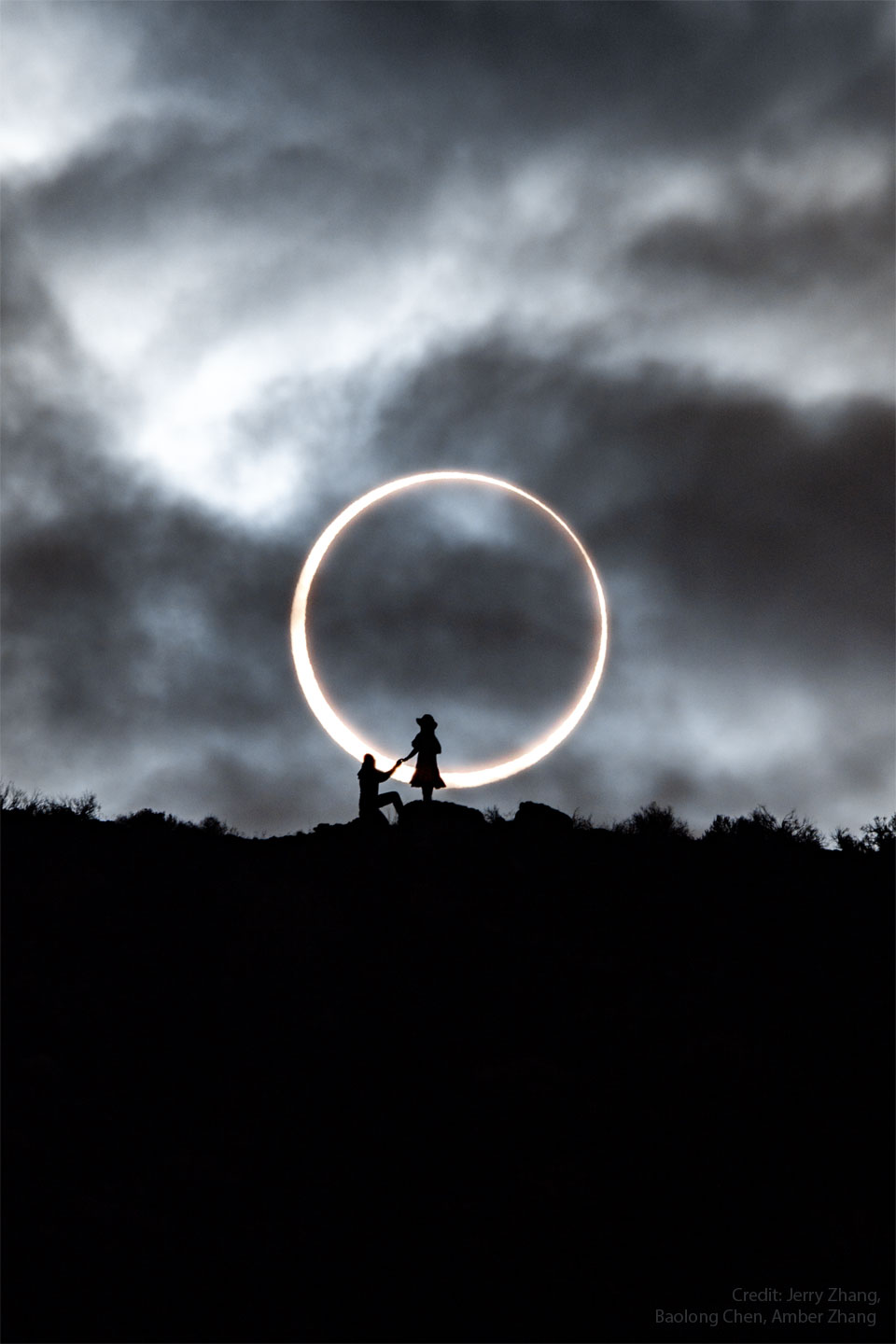 Vor einem dramatisch bewölkten Himmel zeichnen sich am Horizont zwei Personen ab, ein Mann kniet vor einer Frau, beide reichen einander die Hand. Hinter den beiden ist ein riesiger Feuerring zu sehen.