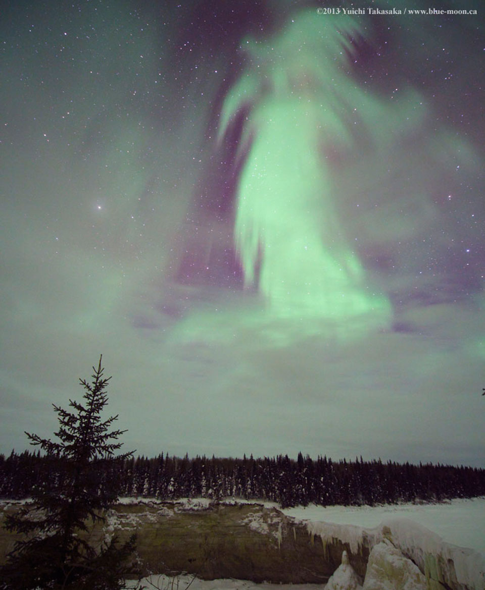 Über einer verschneiten Landschaft mit Wald und Bäumen leuchtet am Himmel ein grünes Polarlicht, dessen Form an einen Geist erinnert.