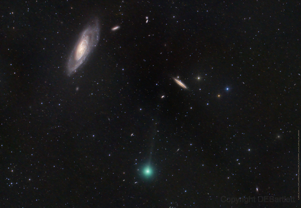Unten in der Mitte leuchtet ein Komet mit grünlicher Koma und einem dünnen, blassen Schweif. Links oben ist eine Galaxie schräg von der Seite zu sehen, rechts über der Mitte sind mehrere kleine Galaxien verteilt.