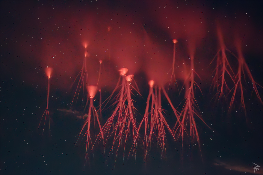 Vor einem dunklen Hintergrund sind Rote Kobolde zu sehen. Aus rötlich leuchtenden trichterförmigen Formen schießen wurzelartige Blitze herab, darüber sind rötlich leuchtende Nebel.