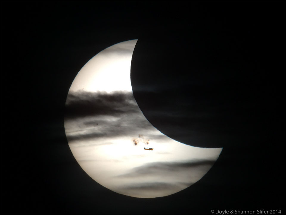 Die Sonne ist teilweise bedeckt von einem Flugzeug, Wolken, dem Mond und Sonnenflecken. Zu sehen ist eine breite Sonnensichel mit dunklen Strukturen.