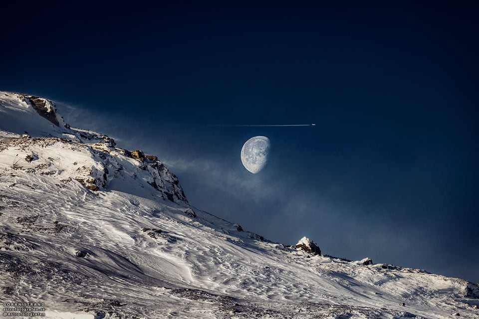 Hinter einem verschneiten Berghang leuchtet der Mond am blauen Himmel, darüber fliegt ein Flugzeug.