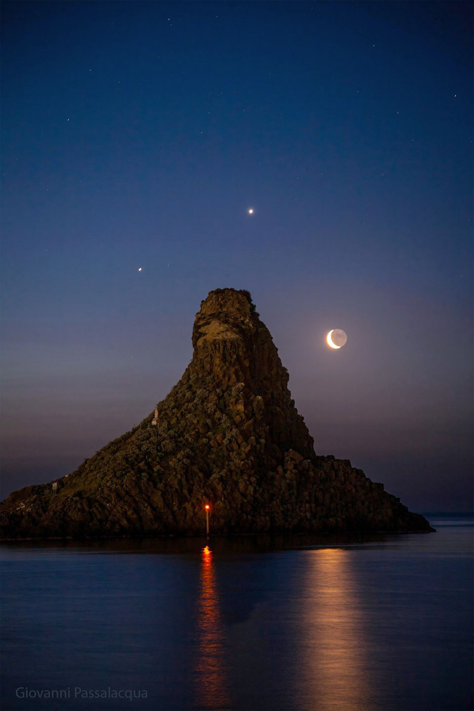 Eine Meereslandschaft umgibt einen großen, mit Bäumen bewachsenen Hügel. Um den Hügel herum sind am Nachthimmel drei helle Punkte zu sehen: die Planeten Jupiter und Venus sowie eine Mondsichel.