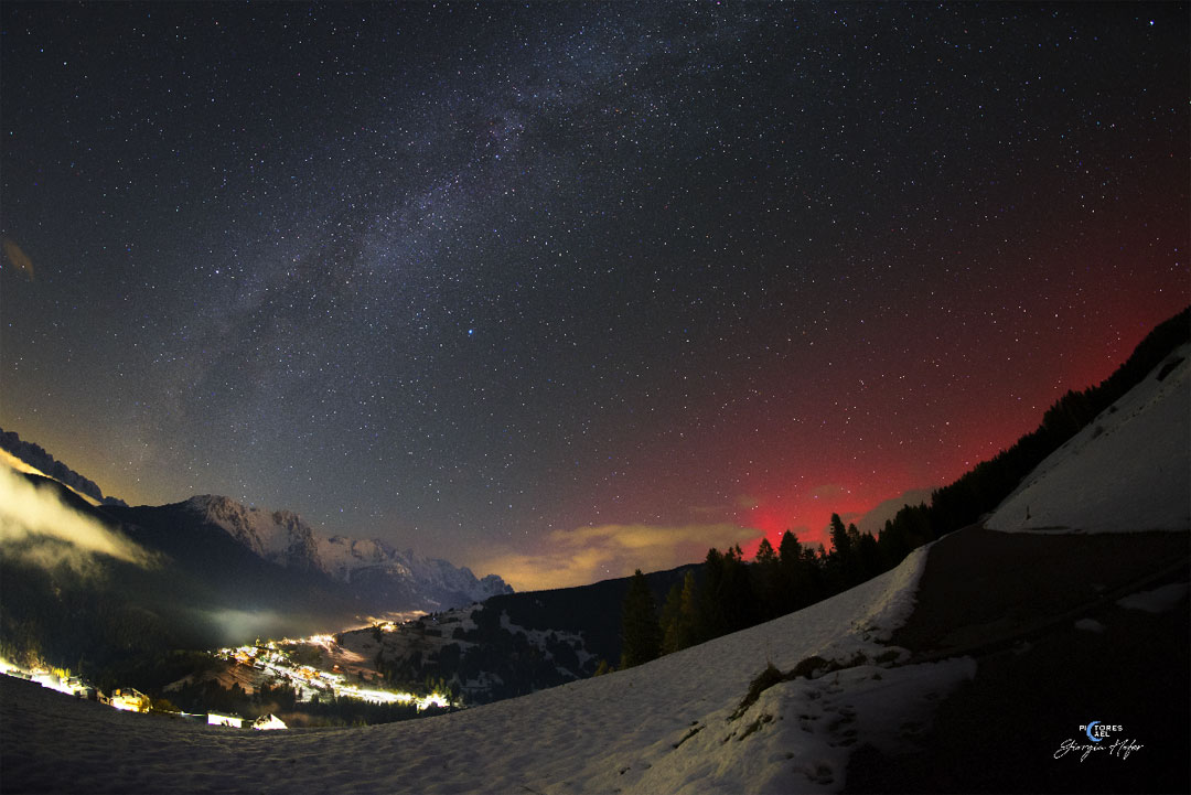 Hinter einem verschneiten Tal mit einer weiter entfernten, lichtdurchfluteten Ansiedlung leuchtet über dem Horizont mit Bergen ein rotes Polarlicht. Oben wölbt sich die Milchstraße.