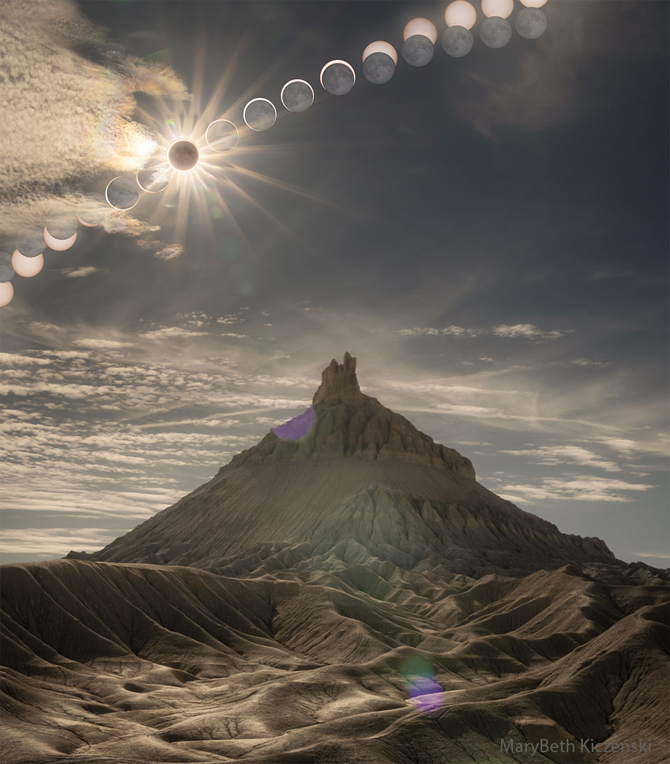 Im Vordergrund ragt hinter einer felsigen Landschaft ein pyramidenförmiger Berg auf. am dramatischen Himmel verläuft von links nach oben diagonal die Sonnenfinsternis in Phasen, in der Mitte ist die Sonne vom Mond bedeckt und von einem Strahlenkranz umgeben.
