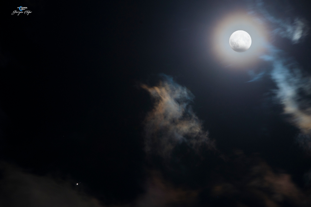Rechts oben leuchtet der Vollmond, am unteren Rand ist ein Teil vom Erdschatten bedeckt, er ist von einem irisierenden Nebelring umgeben. Links unten leuchtet der Planet Jupiter.