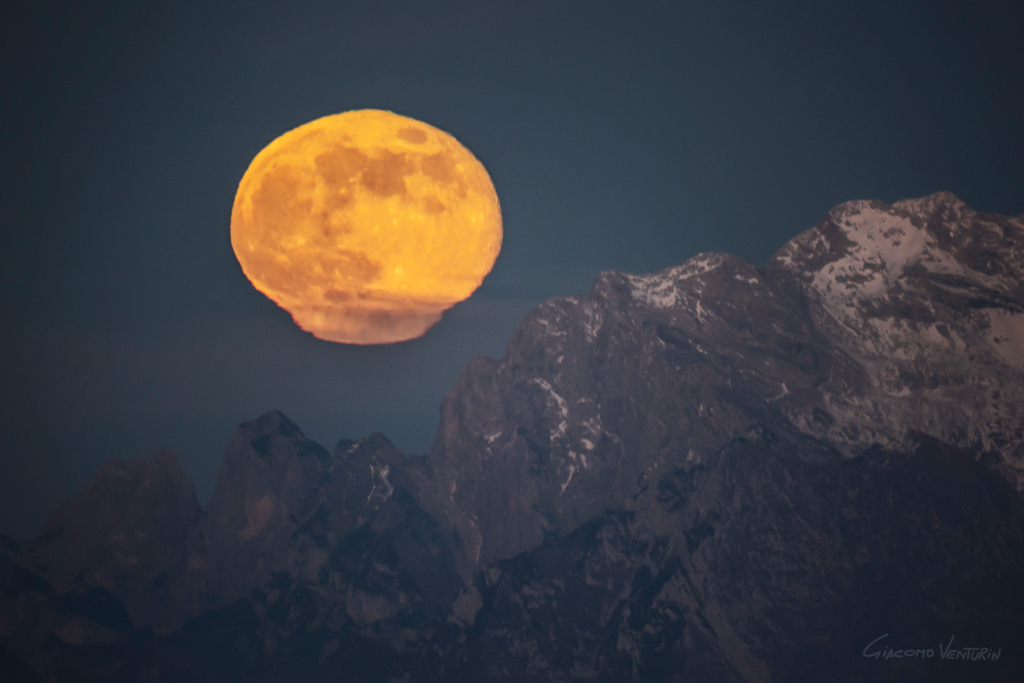 Über einem Gebirge ragt ein gelblicher Mond mit dunklen Meeren auf, er wirkt durch die Atmosphäre über dem Horizont verzerrt. Unten sind Wolken vor dem Mond.