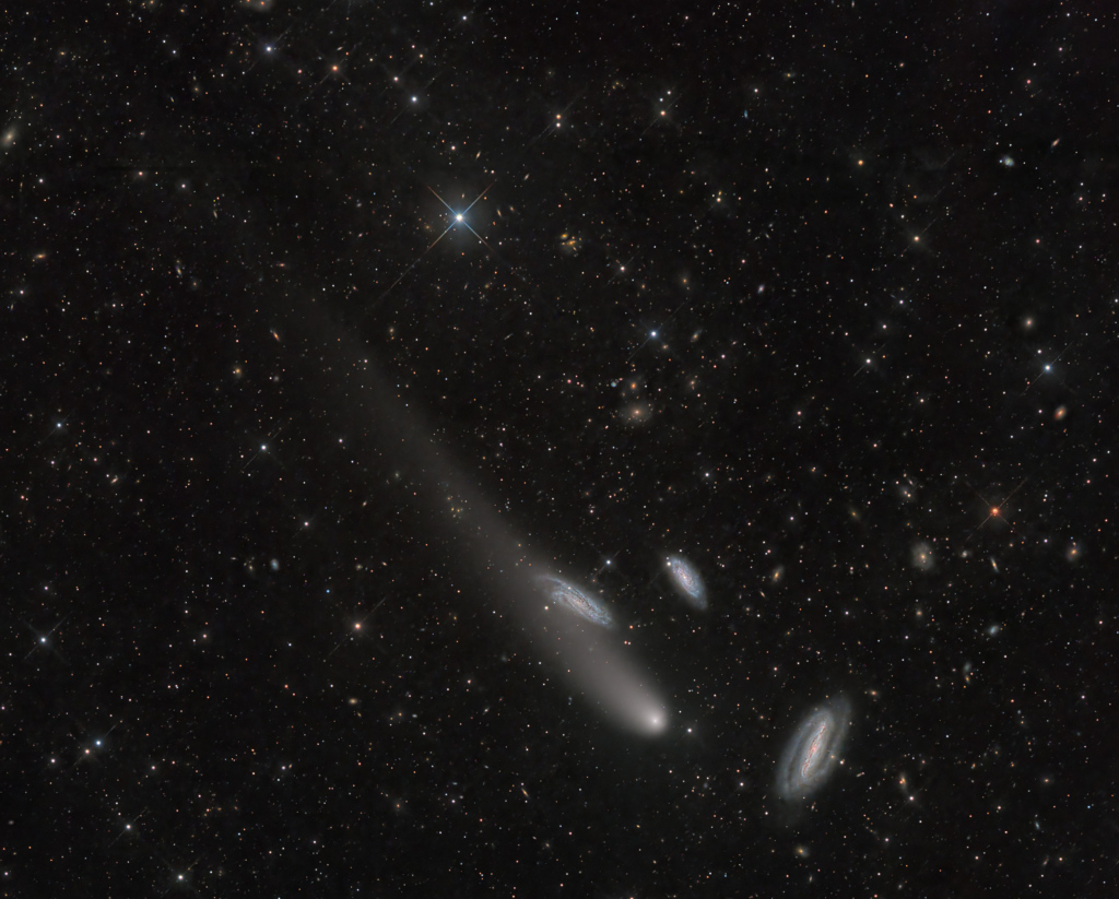 Rechts unten in einem Bildfeld voller Galaxien ist eine Gruppe, die aus einem Kometen und drei Galaxien besteht. Die Galaxien sind schräg von der Seite zu sehen.