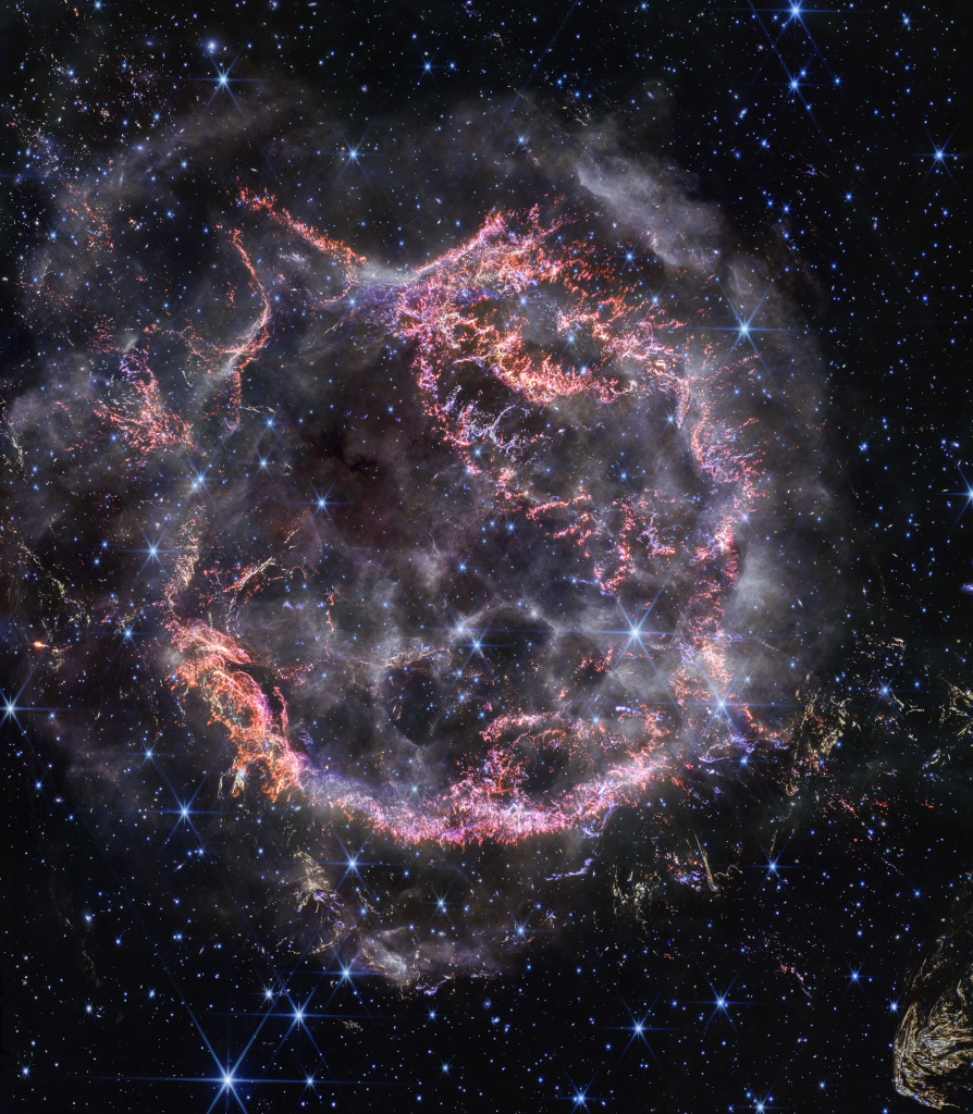 Mitten im Bild prangt eine runde Struktur aus vielen rosa-lila Fasern, die bei einer gewaltigen Sternexplosion entstanden sind. Die Struktur dehnt sich aus. Über allem liegen nebelartige weiße Dunstwolken.