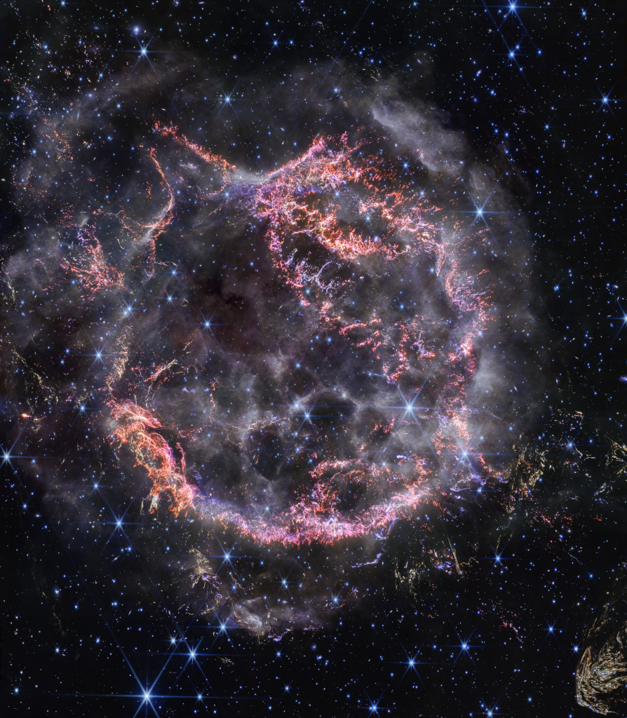 Mitten im Bild prangt eine runde Struktur aus vielen rosa-lila Fasern, die bei einer gewaltigen Sternexplosion entstanden sind. Die Struktur dehnt sich aus. Über allem liegen nebelartige weiße Dunstwolken.