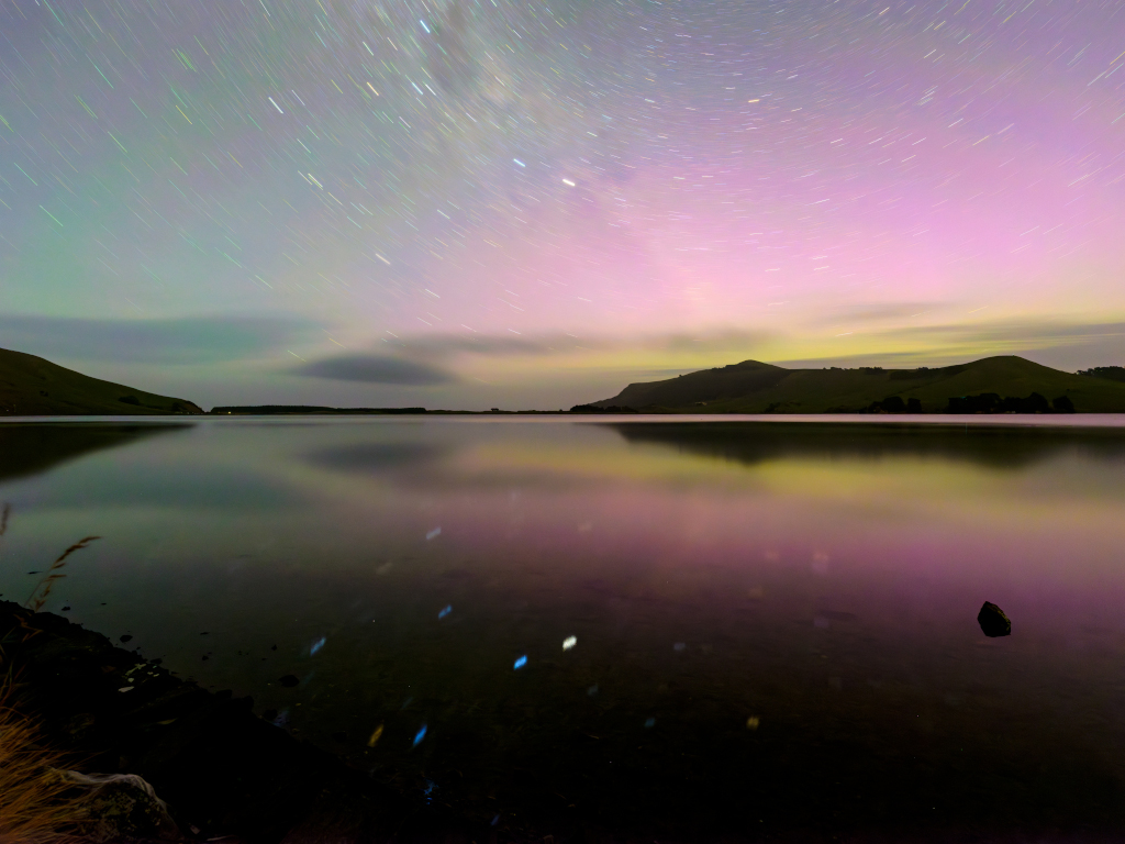 Über einem ruhigen Wasser leuchtet ein Südlicht, der Himmel ist von Strichspuren und schillernden Farben überzogen, am Horizont sind die Silhouetten von Bergen.
