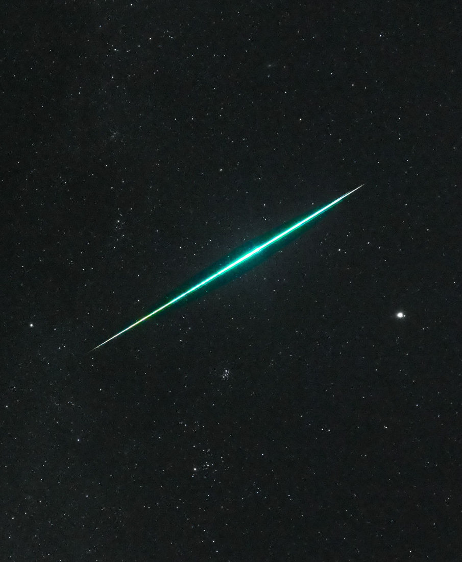 Ein gleißend heller, grün-türkiser Meteor flitzt schräg durch das Bildfeld, im Hintergrund leuchten zarte Sterne, unter der Feuerkugel sind die Plejaden und die Hyaden erkennbar, rechts leuchtet der helle Planet Jupiter.
