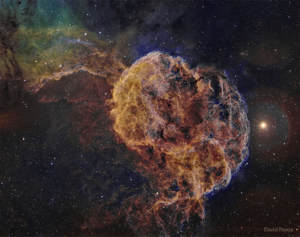 In der Mitte leuchtet ein stark gefaserter, detailreicher quallenförmiger Nebel abgebildet, im Hintergrund sind Sterne und einige weitere Nebel verteilt.