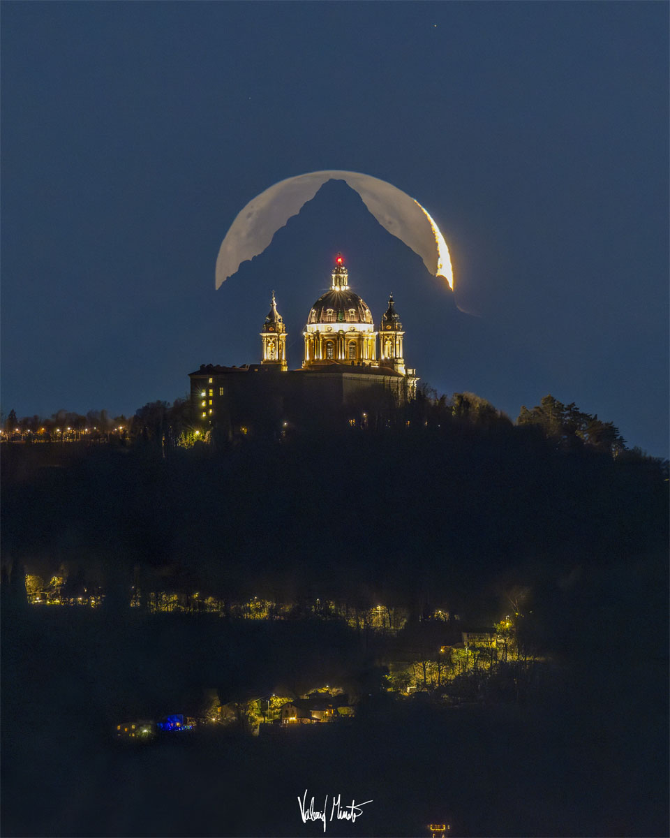 Auf einem Berg steht eine beleuchtete Kathedrale, dahinter ragt ein Berg auf, hinter dem der Mond aufgeht.