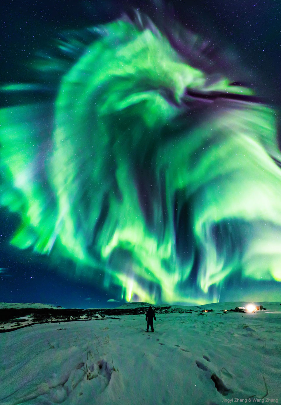 In einer verschneiten, grün beleuchteten Landschaft steht in der Mitte in einiger Entfernung eine Person. Am Himmel strahlt ein Polarlicht, das an einen Drachen erinnert.