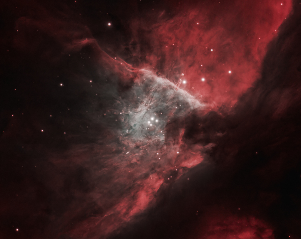 Mitten im Bild ist ein gräulicher Nebel mit einem Sternentrapez, nach rechts oben breitet sich ein rot leuchtender Nebelstreifen aus, auch nach unten reicht ein kleinerer rötlicher Nebelteil.