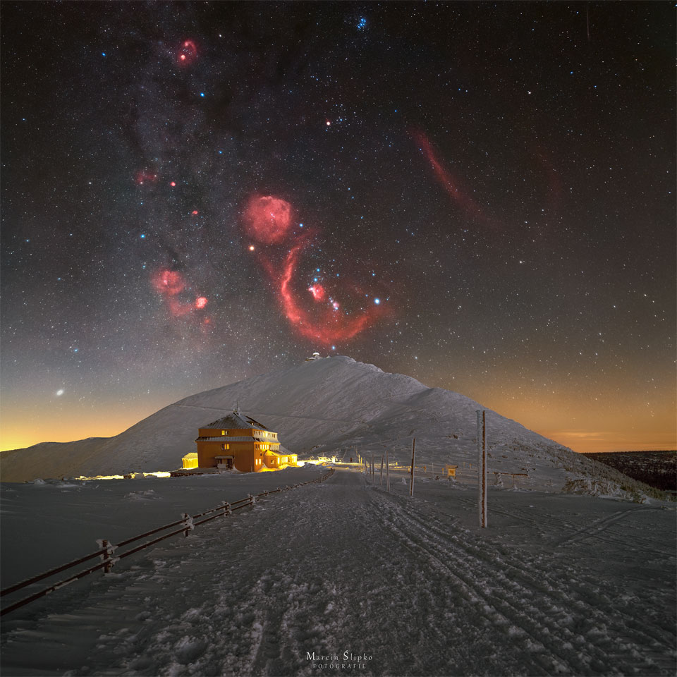 Das Bild zeigt eine verschneite Landschaft mit einem großen Hügel in der Mitte. Über dem Hügel sind die Sterne und Nebel des Sternbildes Orion zu sehen. Das rote Leuchten der Nebel steht in starkem Kontrast zum dunklen Himmel und dem hellen Schnee.