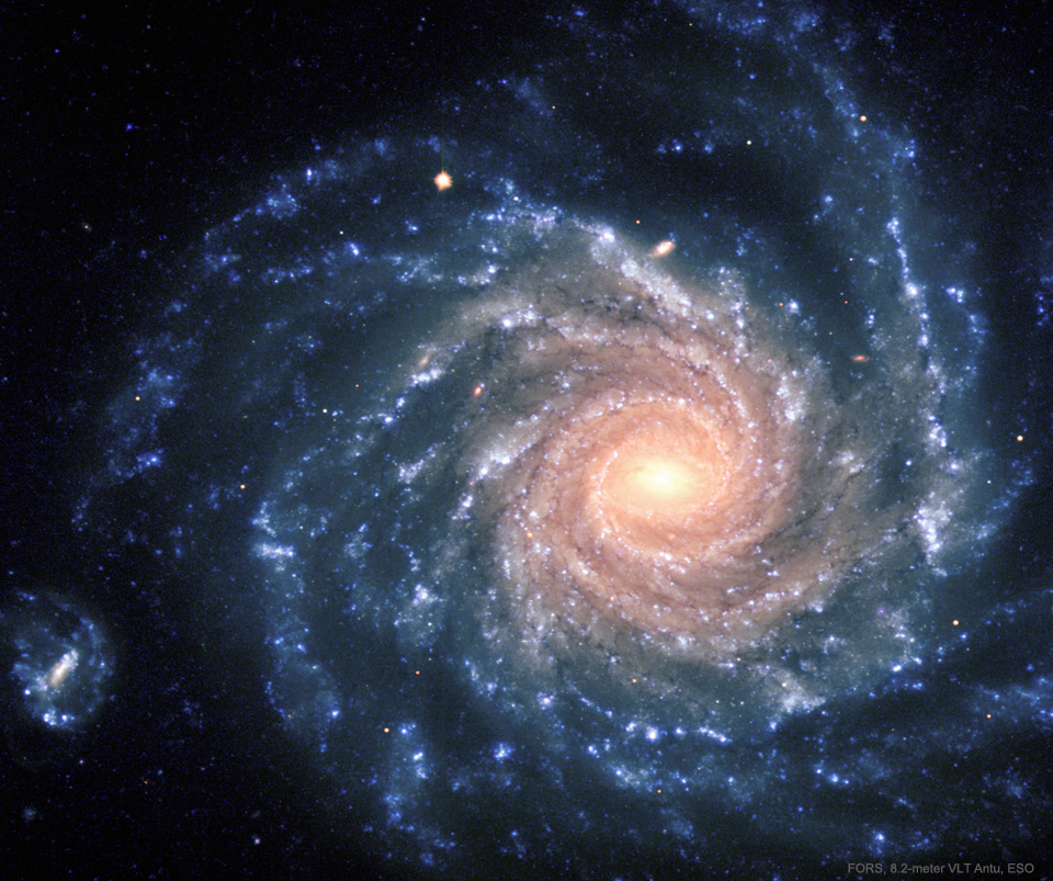 Bildfüllend ist eine Spiralgalaxie dargestellt, die wir direkt von oben sehen. In der Mitte leuchtet ein hellgelbes Zentrum, außen herum verlaufen stark strukturierte Spiralarme. Am rechten Bildrand ist eine kleine Galaxie.