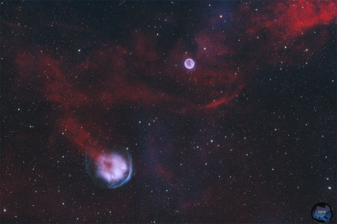 Zwischen roten Nebeln und wenigen Sternen leuchten zwei helle lila runde Nebel.