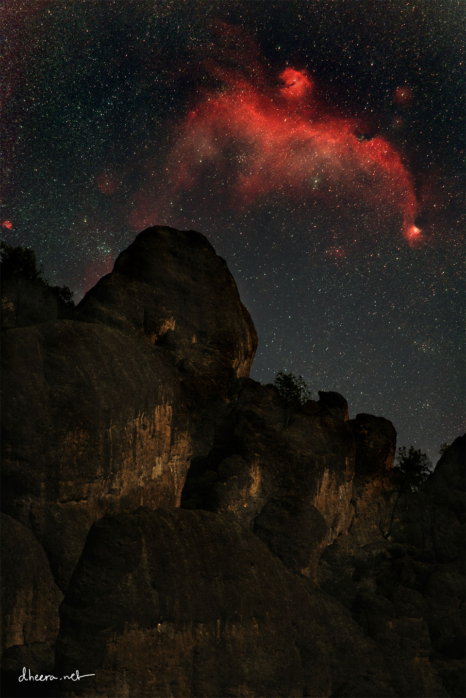 Über der dunklen Silhouette eines Berges mit einem markanten Gipfel schwebt am sternklaren Himmel ein roter Nebel, der an eine Möwe erinnert, der Möwennebel.