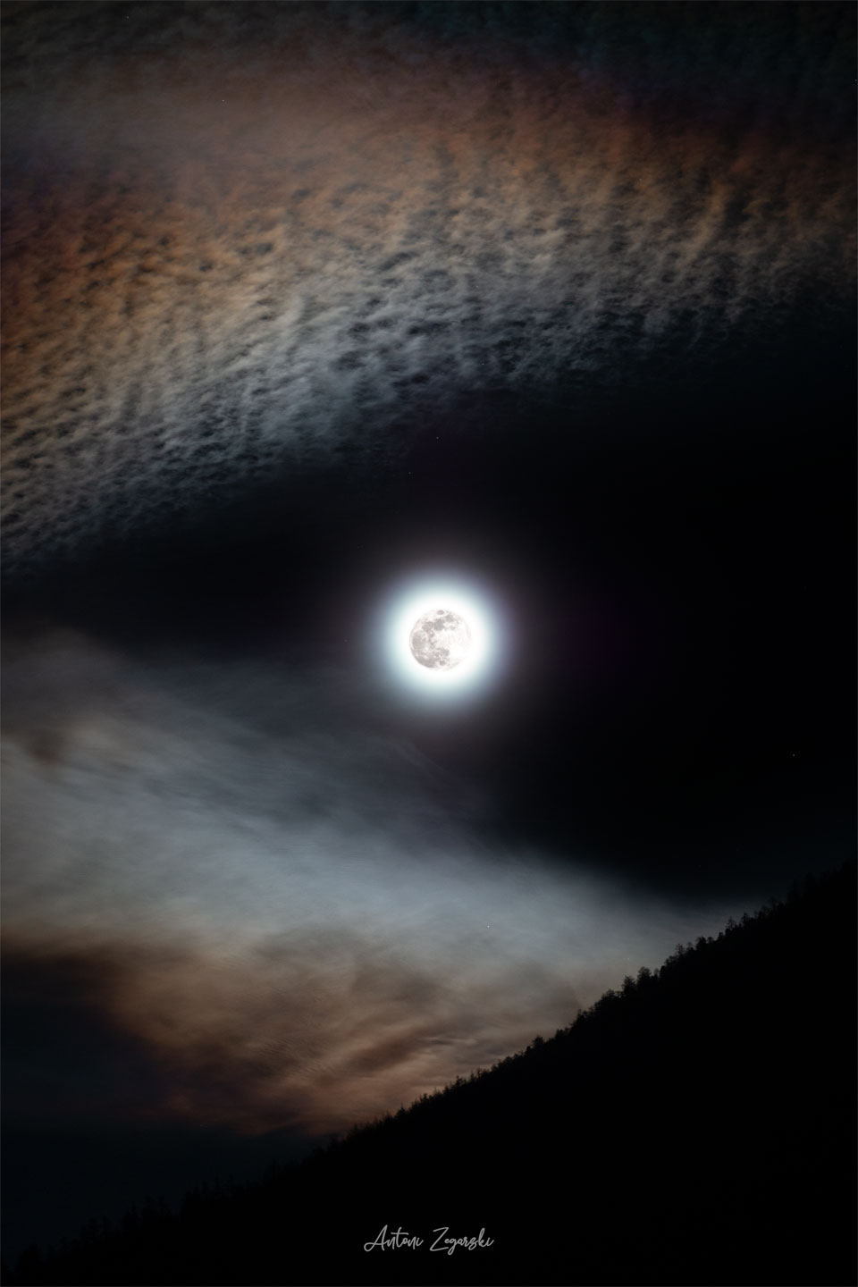 Mitten im Bild leuchtet der Mond, umgeben von einem hellen Lichterkranz. Oben sind regenbogenfarbige Zirren, unten ein dunkler bewaldeter Berghang.