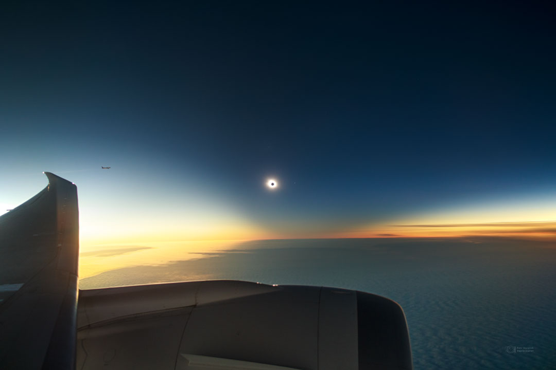 Über dem Flügel eines Flugzeuges ist ein klarer, abgeschatteter dunkelblauer Himmel, in der Mitte leuchtet die Sonnenkorona rund um den schwarzen Mond. Links und rechts neben dem Schatten ist der Himmel hellorange gefärbt.