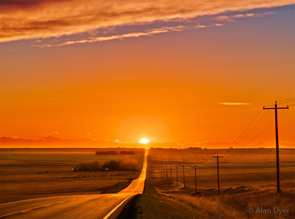 Eine Straße führt in den Sonnenuntergang. Die Landschaft ist leicht wellig, der Blick reicht sehr weit zum Sonnenuntergang. Der Himmel ist klar, die ganze Landschaft ist in ein tiefes Orange getaucht.
