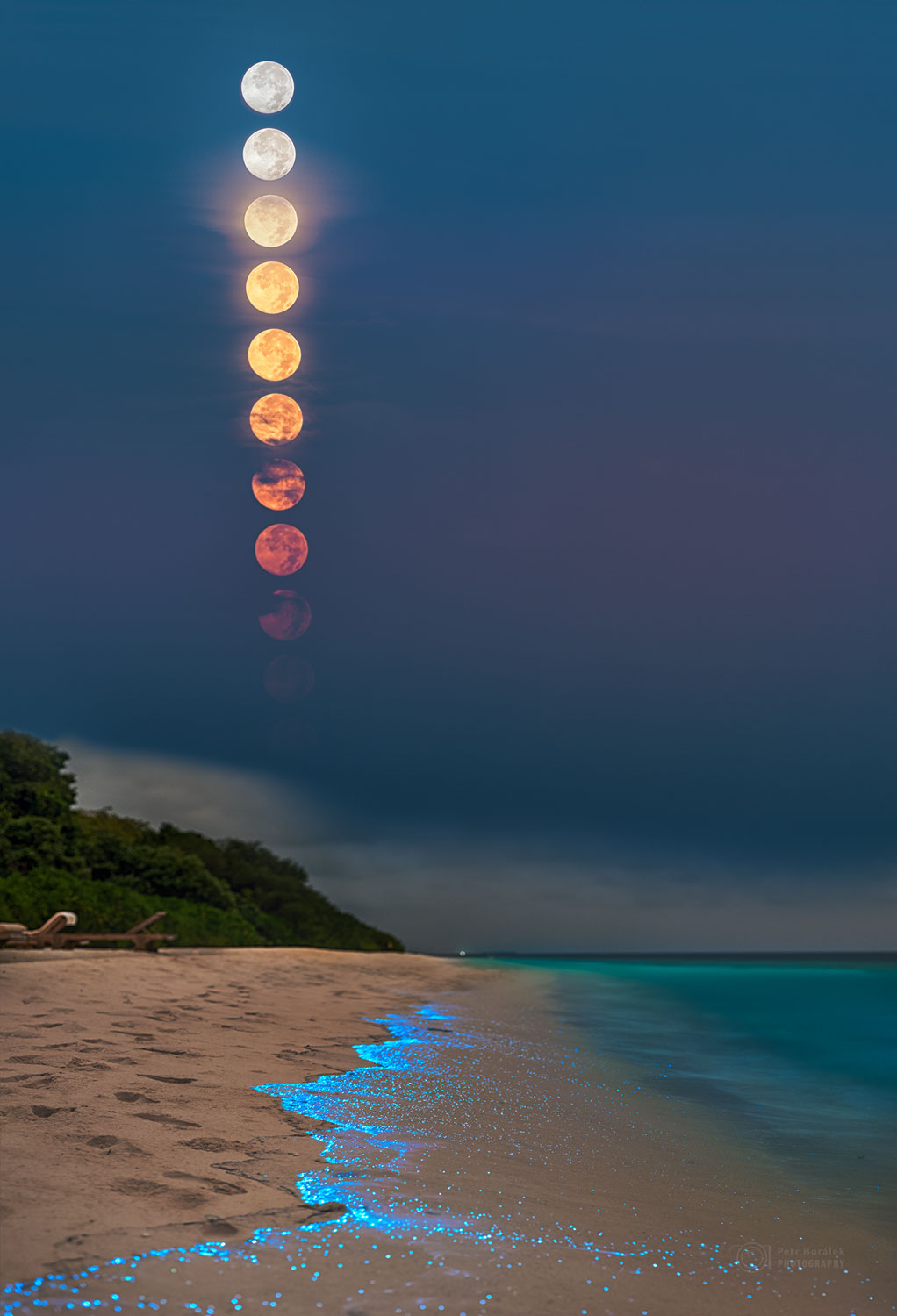 Über einem Strand steigt der Vollmond senkrecht unter. Es sind 8 Bilder des Mondes zu sehen, die untersten sind rötlich, die obersten beiden weißgrau. Das Wasser am Strand hat einen blau leuchtenden Saum.