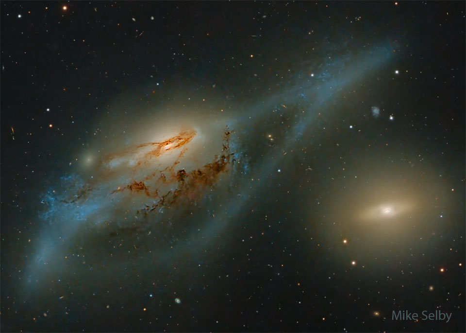 Zwei Galaxien leuchten im Bild, die linke Galaxie ist von einer blauen blassen Schleife aus Sternen umgeben.