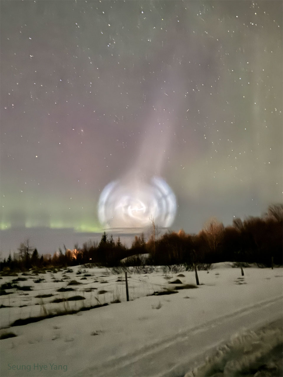 Hinter einem verschneiten Hang verläuft ein Wald am Horizont, dahinter strahlt in der Bildmitte eine weiß leuchtende Spirale, von der ein weniger heller Strahl nach rechts oben ausgeht. Links über dem Horizont sind Polarlichter zu sehen.