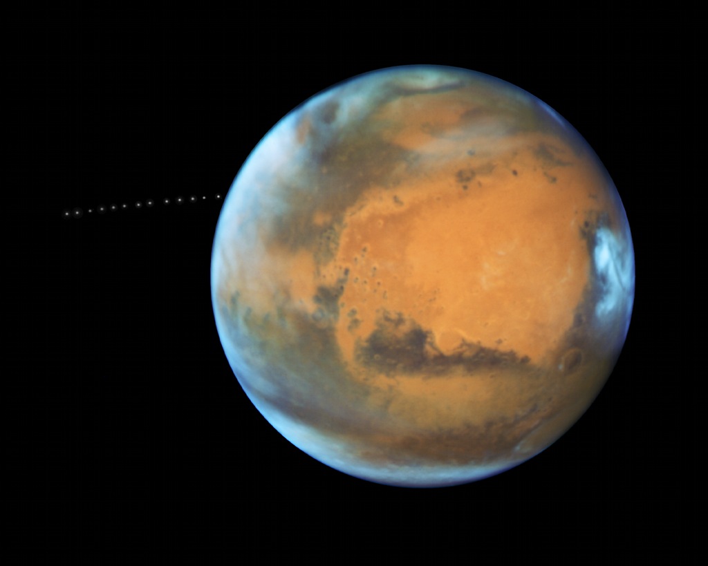 Der Mars schwebt als leuchtende Kugel im Raum, die Polkappe oben ist schwach ausgeprägt, rechts ist ein markanter heller Fleck. Links ist eine Kette aus winzigen Lichtpunkten, es ist der Marsmond Phobos.