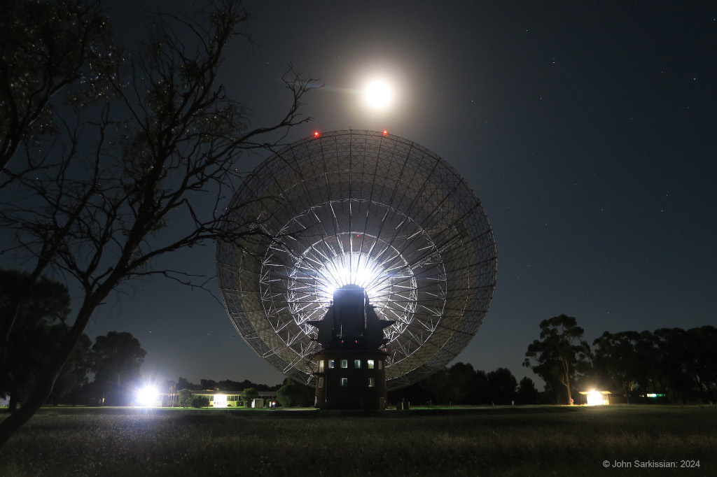 Mitten im Bild steht eine riesige Radioantenne, die in der Mitte beleuchtet ist. Darübr steht der Vollmond. Im Hintergrund am Horizont stehen Bäume.