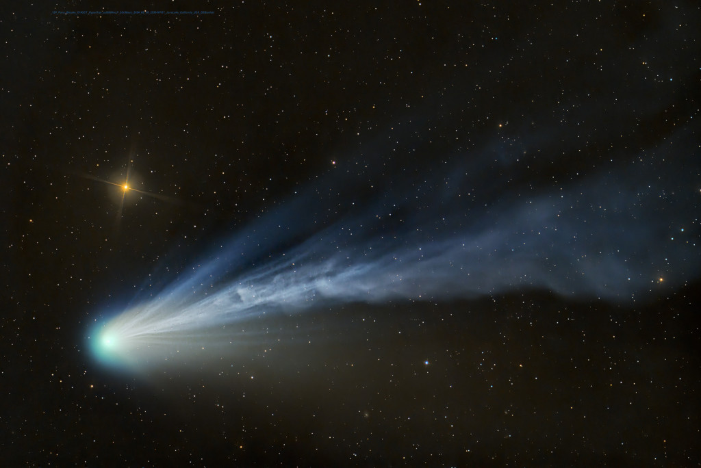 Der Komet im Bild hat links eine grünliche Koma, darüber leuchtet ein orangefarbener Stern mit Zacken. Nach rechts fächern sich die Schweife auf, die meisten davon wirkden strahlenförmig, nur der Schweif in der Mitte erinnert an eine Rauchschwade.