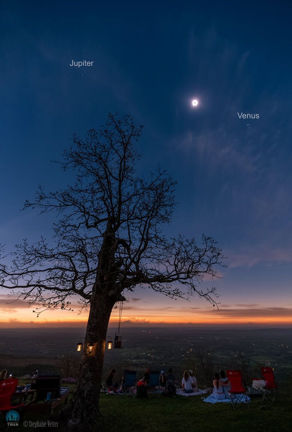 Am Himmel ist eine vollständig verfinsterte Sonne zu sehen, die von einer hellen Korona umgeben ist. Im Vordergrund beobachten mehrere Personen die Sonne in der Nähe eines großen Baumes. Rechts neben der verfinsterten Sonne ist der helle Planet Venus zu sehen, links daneben der fast gleich helle Planet Jupiter.