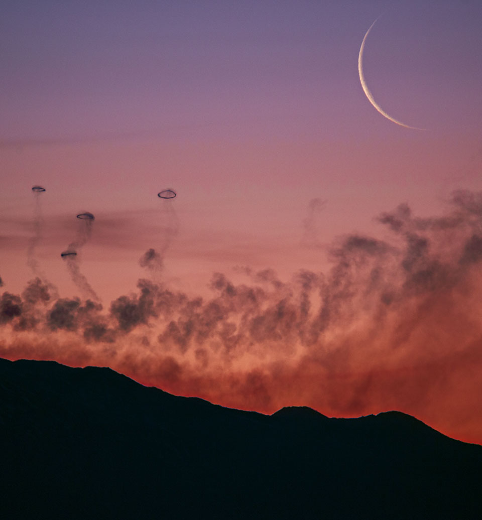 Über der Silhouette eines Berges steigt Rauch auf und bildet Ringe. Nach oben hin wird der rötliche HImmel violett, rechts oben leuchtet eine schmale Mondsichel.