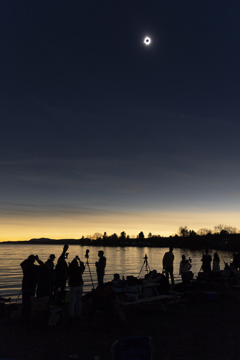 Über einem See mit vielen Schaulustigen strahlt am dunklen Himmel die Korona der Sonne um den Mond herum. Am Horizont ist der Himmel hell.