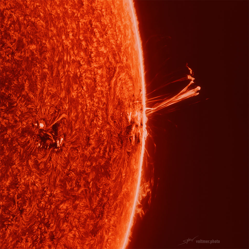 Die linke Bildhälfte füllt ein Teil der Sonne, durch die Mitte verläuft senkrecht der helle Rand. Nach rechts ragen schleifenförmige Protuberanzen in den schwarzen Weltraum.
