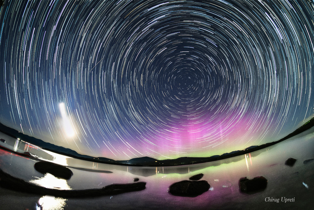 Über einem Stausee und Hügeln leuchten am Himmel Strichspuren und ein leuchtendes Polarlicht, unten grün, oben magentafarben. Links leuchtet der zunehmende Sichelmond und spiegelt sich im Wasser.