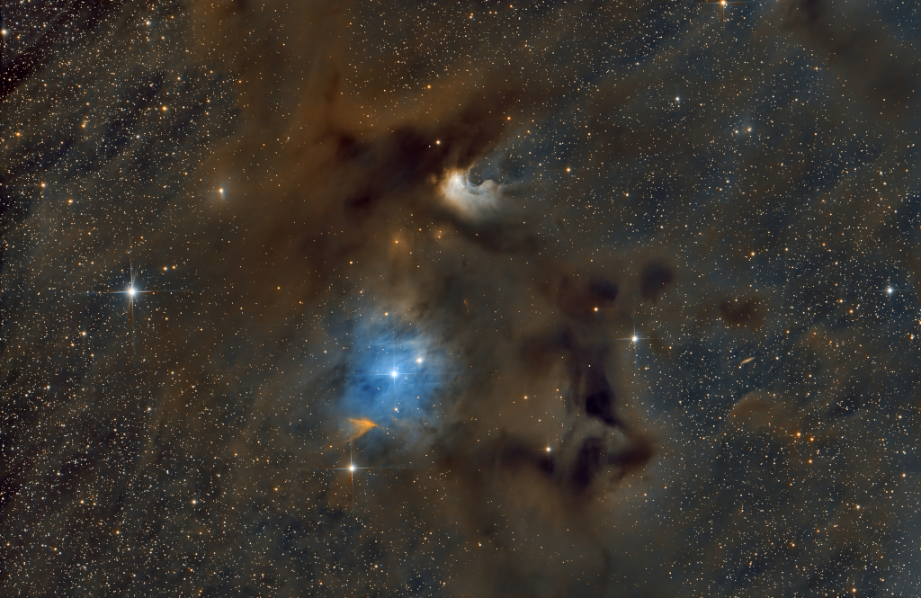 In einem braunen, undurchsichtigen Nebel leuchtet ein blauer Reflexionsnebel mit einem Stern in der Mitte. Links unter dem blauen Nebel ist ein orangefarbener trichterförmiger Nebel. Außen sind kleine Sterne sehr dicht verteilt.