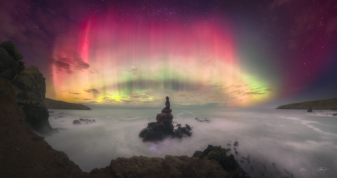 Hinter der Küste über einem wogenden Meer, aus dem ein Steinturm herausragt, leuchtet über dem Horizont ein Polarlicht. Es ist senkrecht gestreift, unten verläuft ein gelbgrüner Bogen, oben leuchten magentafarbene Strahlen.